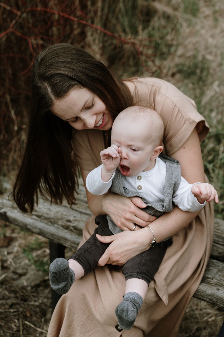 A tender moment between parent and baby sitting outdoors in nature, as the little one yawns or rubs their eyes.