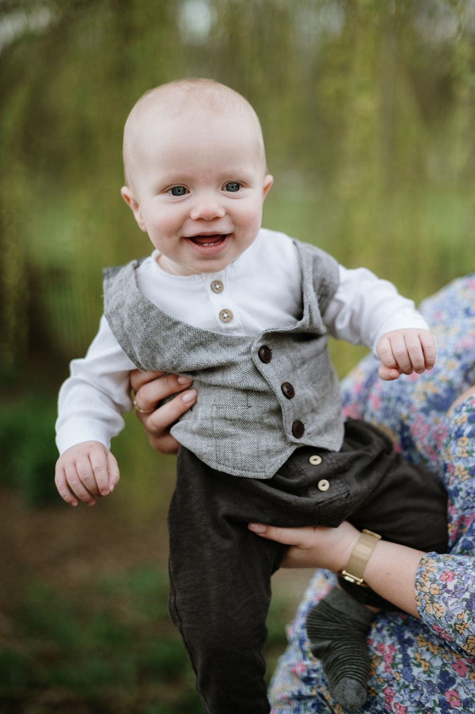 A baby in a gray vest and white shirt smiles while being held outdoors in a natural setting.