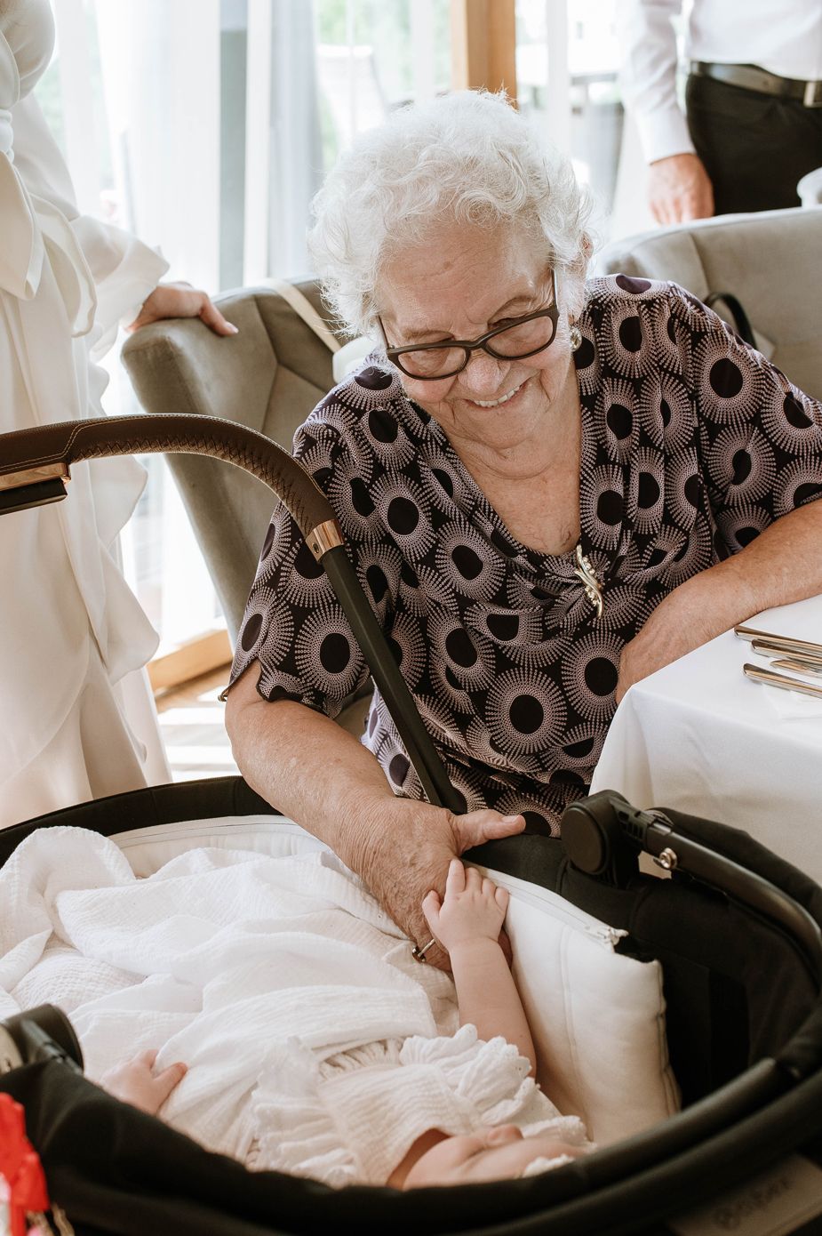 A senior woman wearing glasses and a patterned dress smiles while looking into a baby carrier.