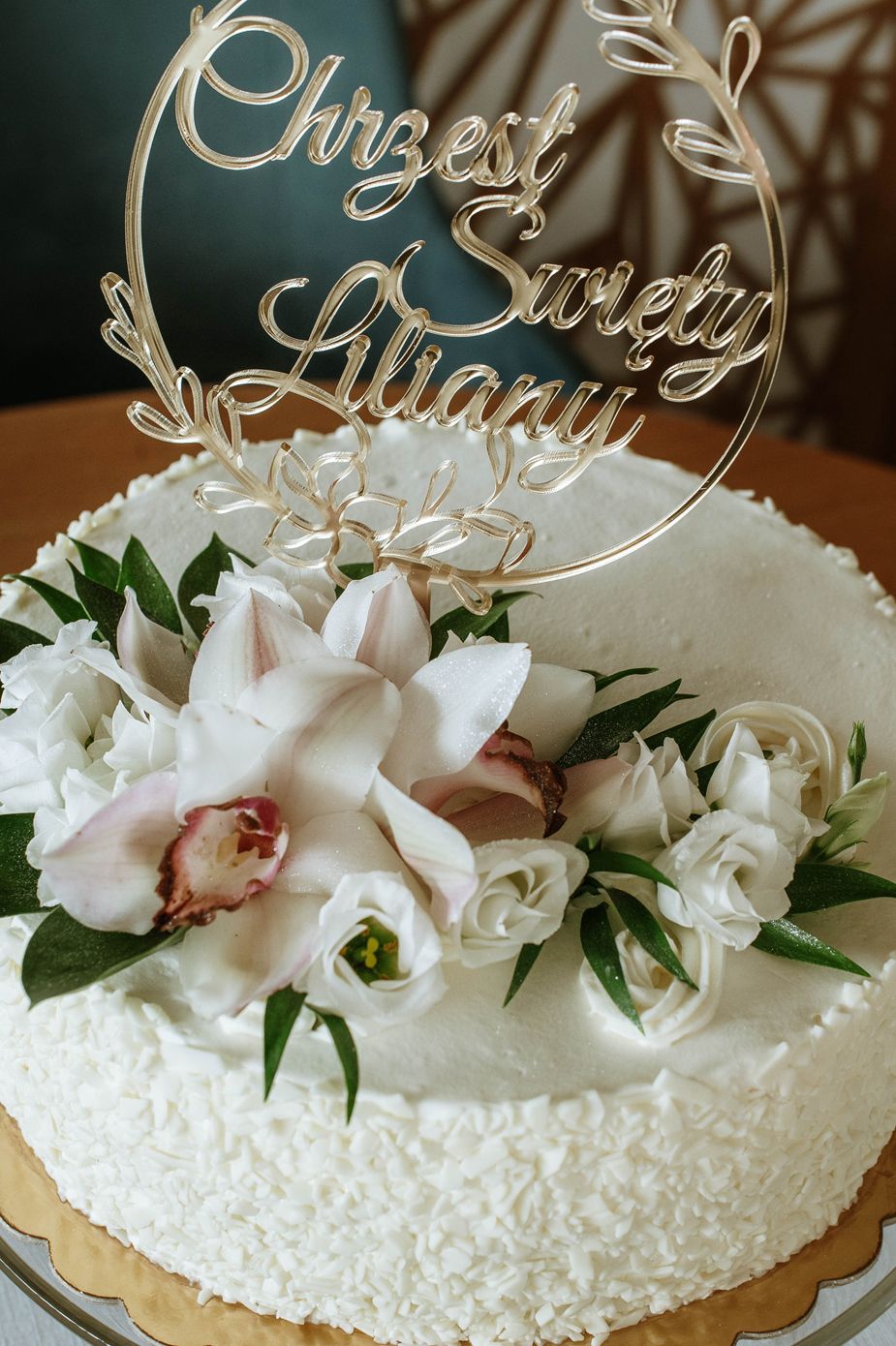 White wedding cake decorated with orchids, roses and a gold circular topper reading 'Our Sweet Always'.