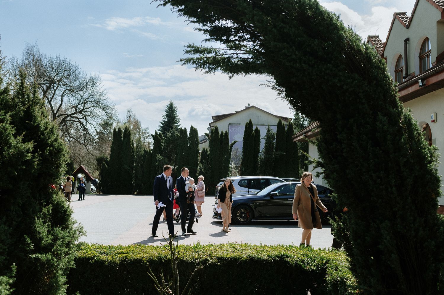 People gathered outside a Mediterranean-style building with cypress trees framing the scene on a sunny day.