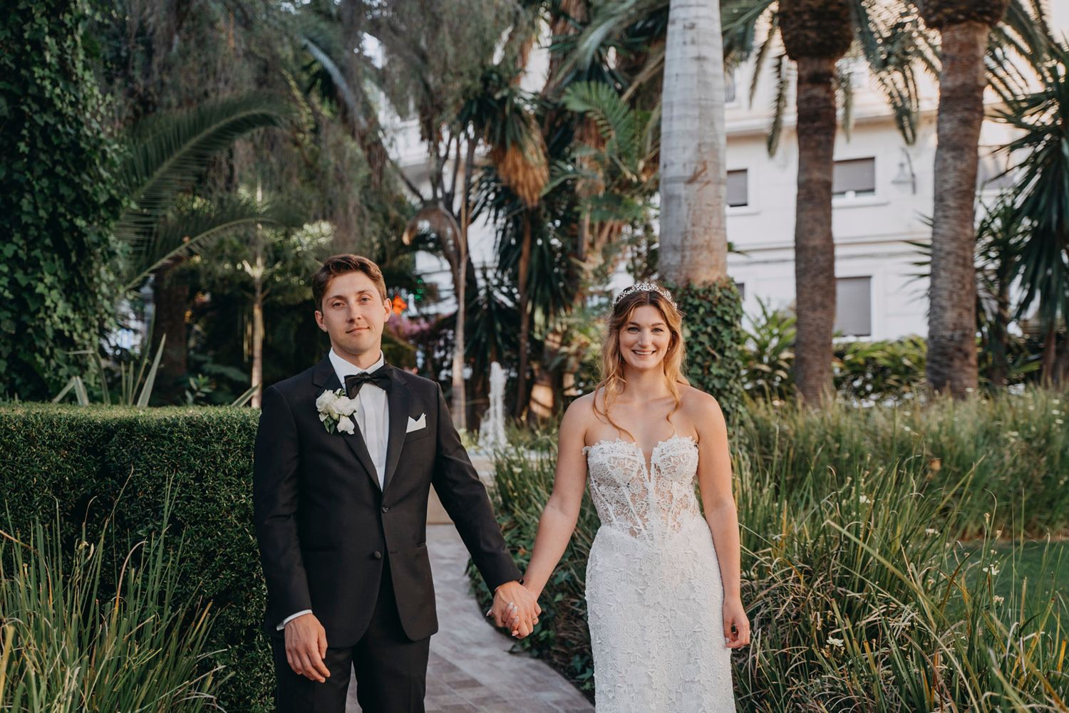 A wedding couple poses together in a lush garden setting with palm trees and natural greenery in the background.