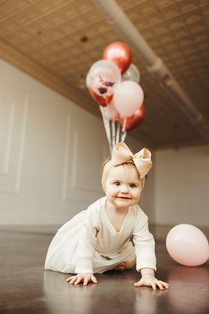 Cheerful baby girl in white tutu dress with bow headband crawls on floor surrounded by pink and rose gold balloons.