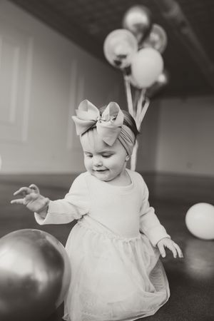 Black and white photo of baby girl with large bow headband playing with metallic balloon at first birthday party.