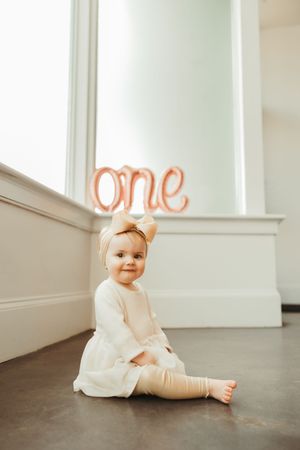 Baby girl in white dress sits on floor smiling, with rose gold 'one' balloon for first birthday.