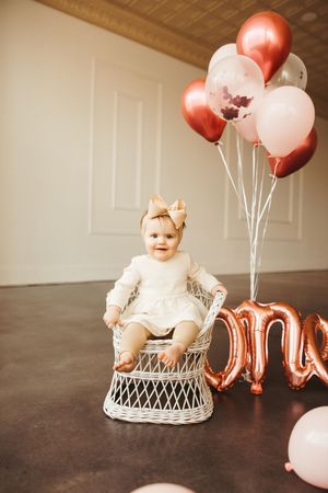 Smiling baby girl in white dress with gold bow headband sits in wicker chair beside rose gold one balloon display.