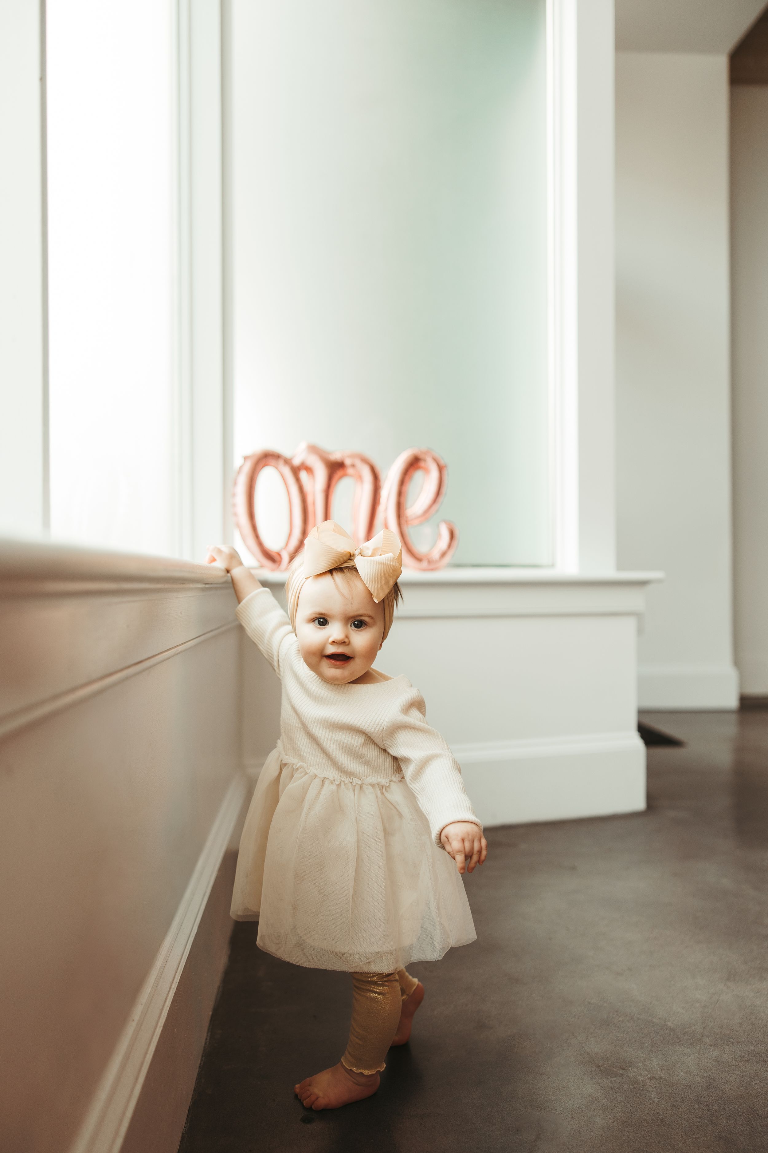 Adorable baby girl in white tutu dress stands holding rose gold 'one' balloon at first birthday.