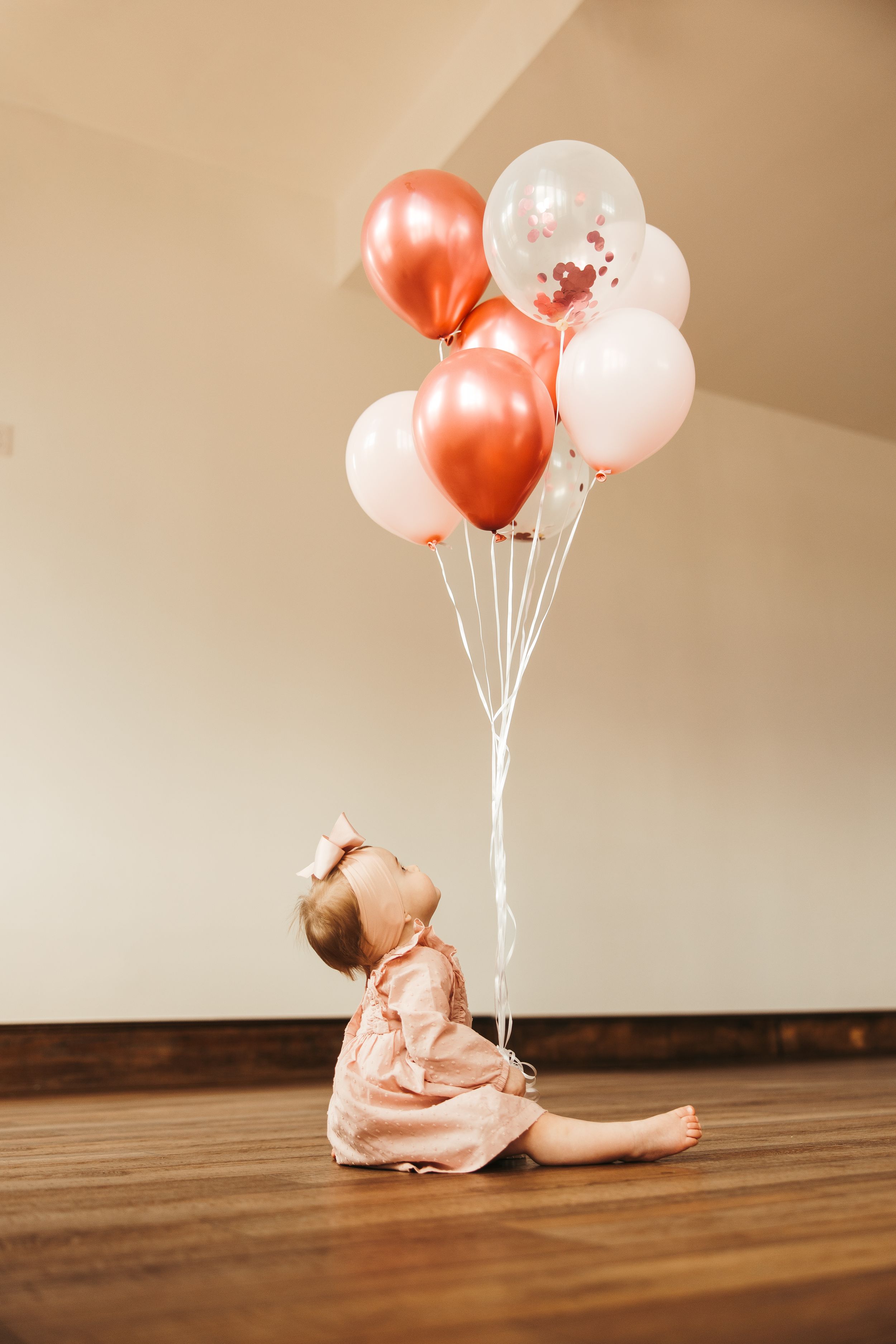 Baby girl in pink dress sits on wood floor gazing up at rose gold and white birthday balloons. First birthday session. Greensboro NC
