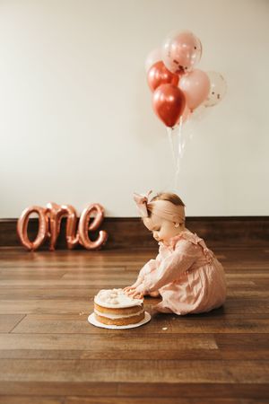 Baby girl in pink dress smashes first birthday cake surrounded by rose gold balloons and 'one' sign.