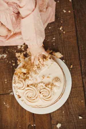 Overhead view of baby's hand smashing white frosted first birthday cake on rustic wood floor.