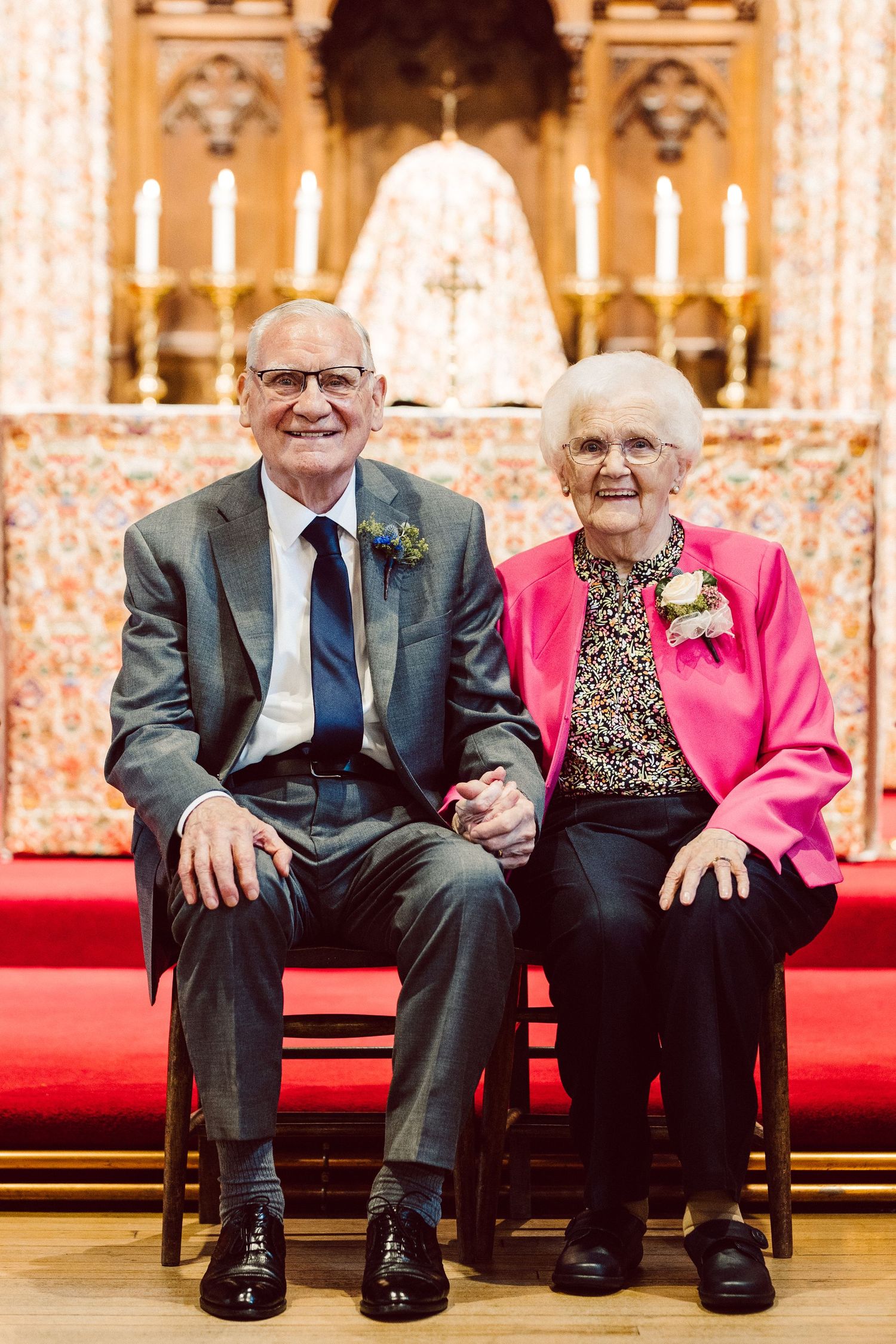 An elderly couple shares a joyful moment while sitting together on red carpet steps in an ornate church setting.