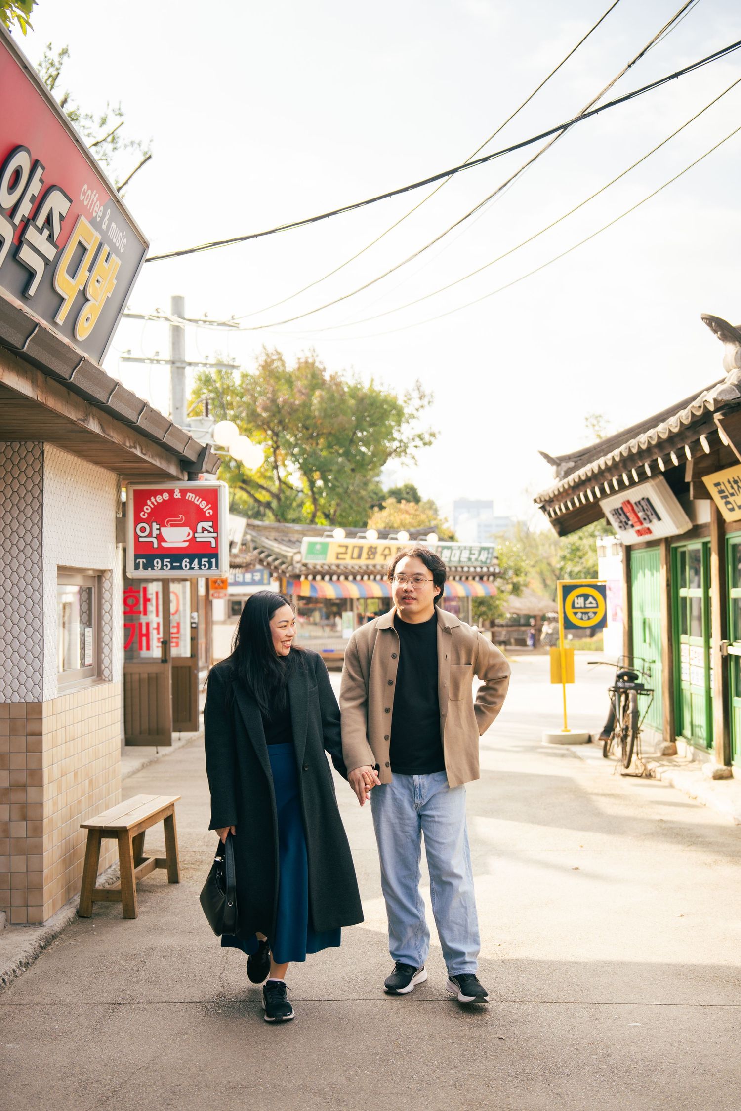 Two people walking together on a street with traditional Asian architecture and power lines overhead.