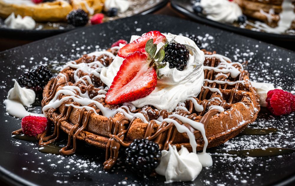 Chocolate waffles topped with whipped cream, strawberries, and powdered sugar on black plates.