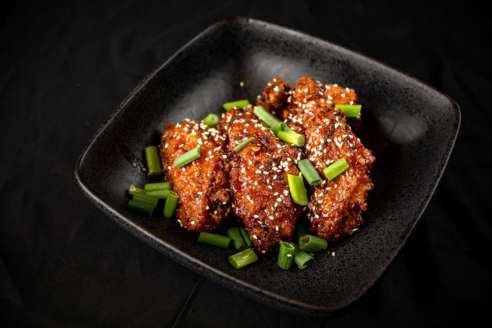 Crispy glazed chicken wings garnished with sesame seeds and green onions on a black plate.