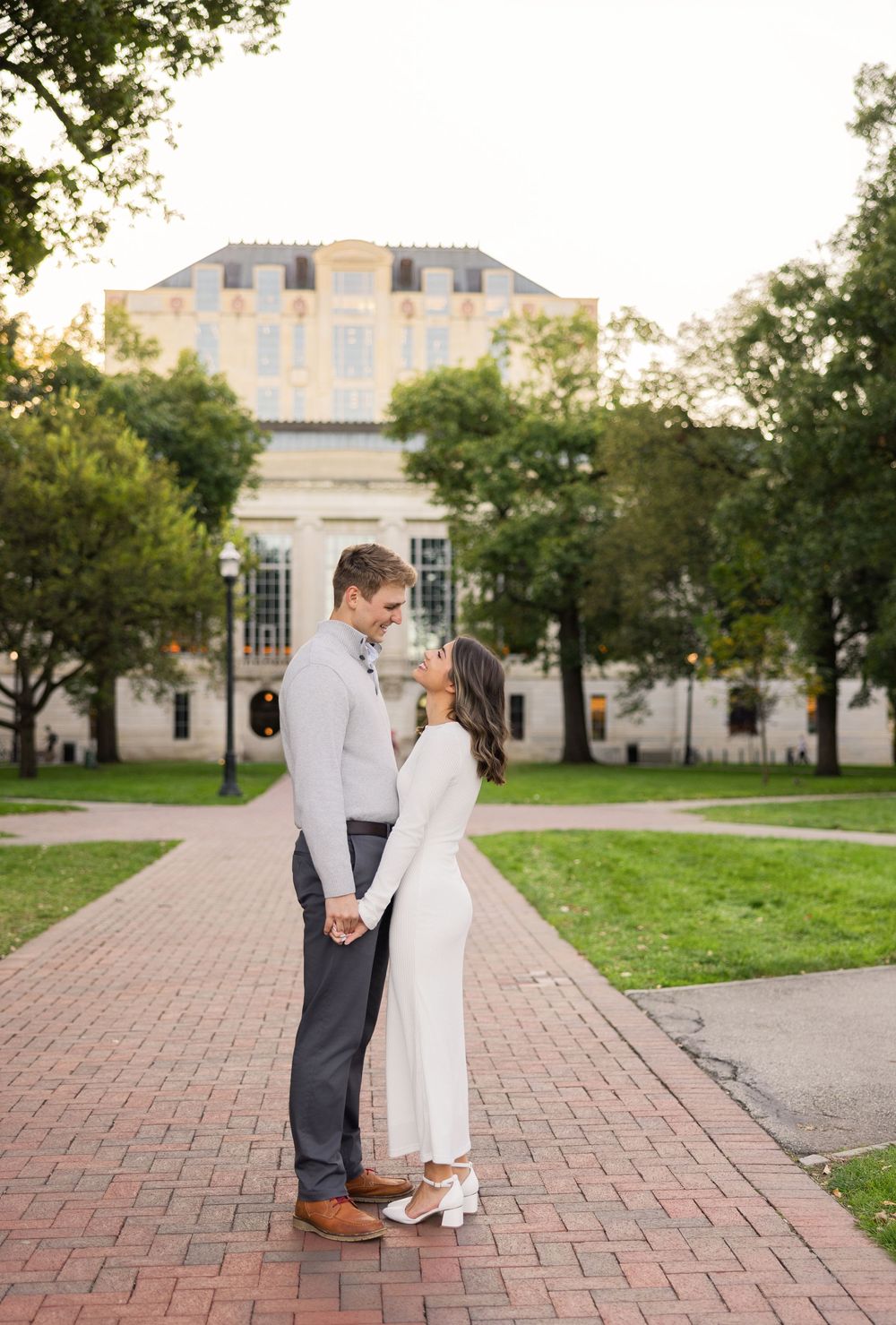 A couple shares a romantic moment on a brick pathway in front of a grand historic building with white columns and architecture.