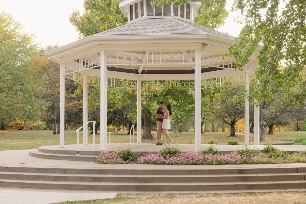 A white gazebo pavilion with curved steps surrounded by landscaped gardens and autumn trees in a park setting.