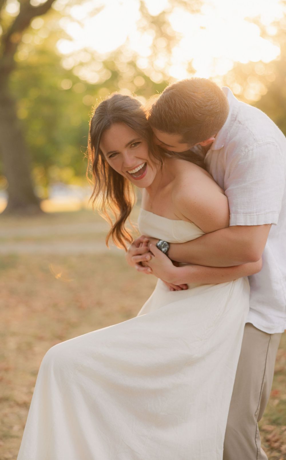 A romantic couple embraces and laughs together during a sunset photoshoot in a park with golden autumn light.