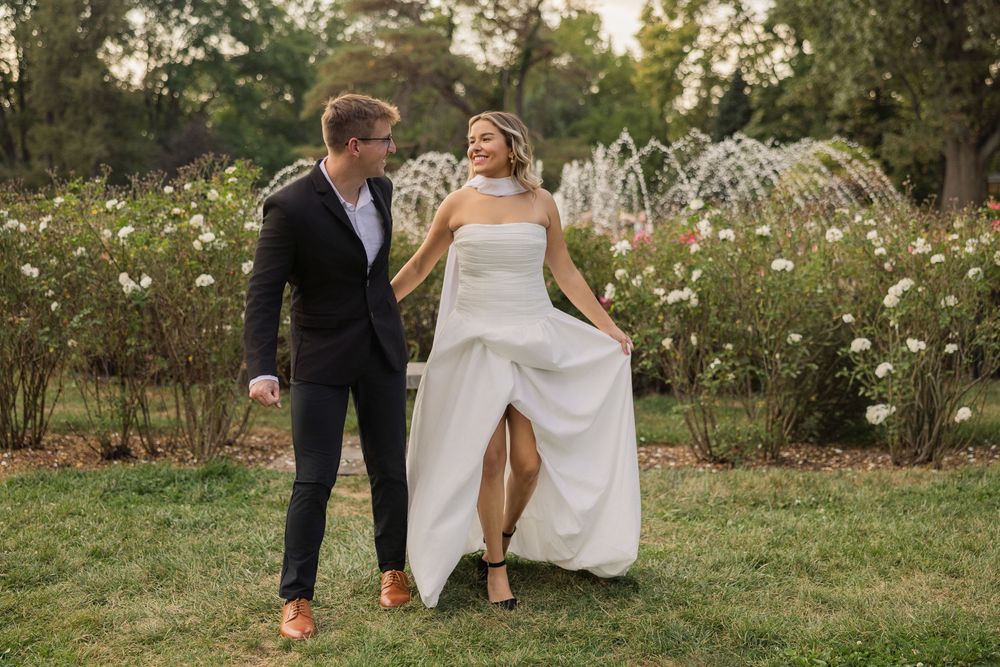 Couple in wedding attire playfully running and dancing through a garden setting at sunset.