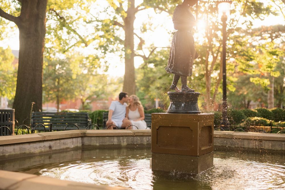 A romantic couple embraces during golden hour sunset in a scenic park setting with autumn foliage in the background.