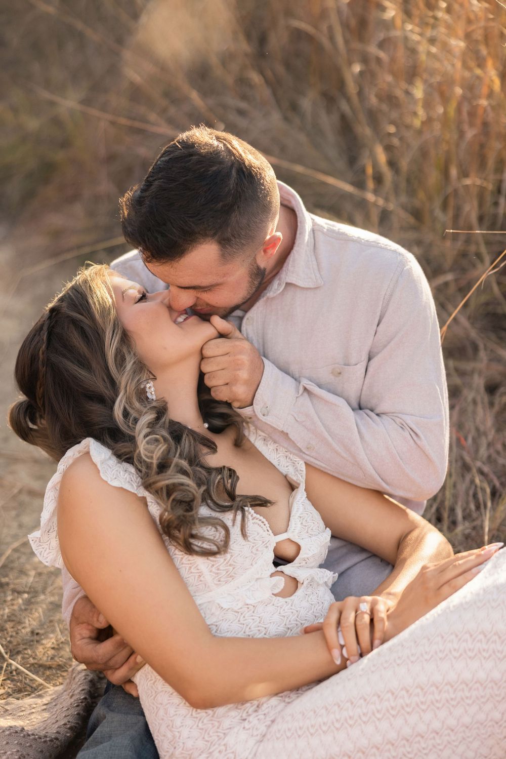 A romantic couple shares tender kisses while lying in golden sunlit grass during an outdoor photo session.