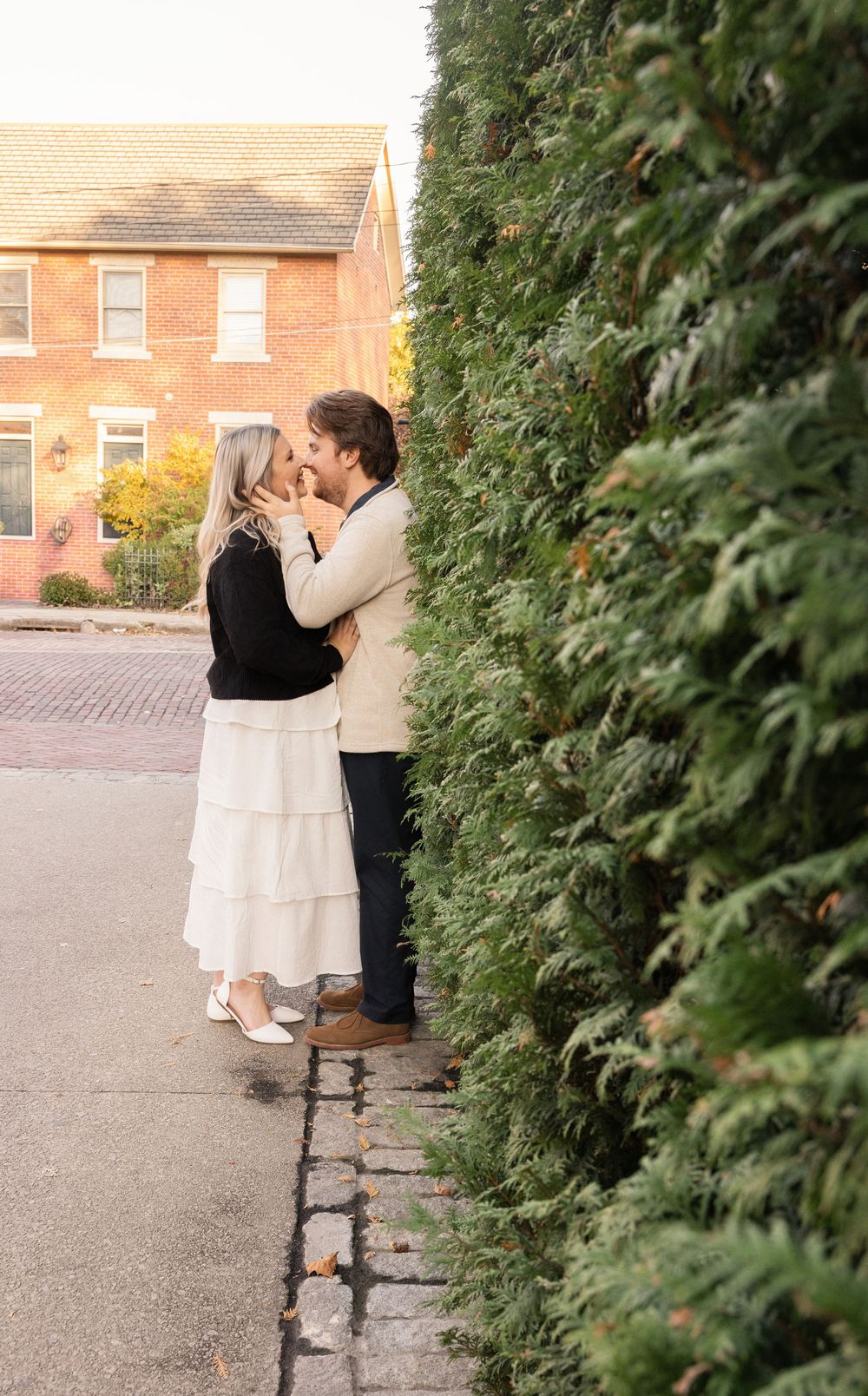 A romantic couple embraces on a brick sidewalk next to evergreen shrubs with a brick house in the background.