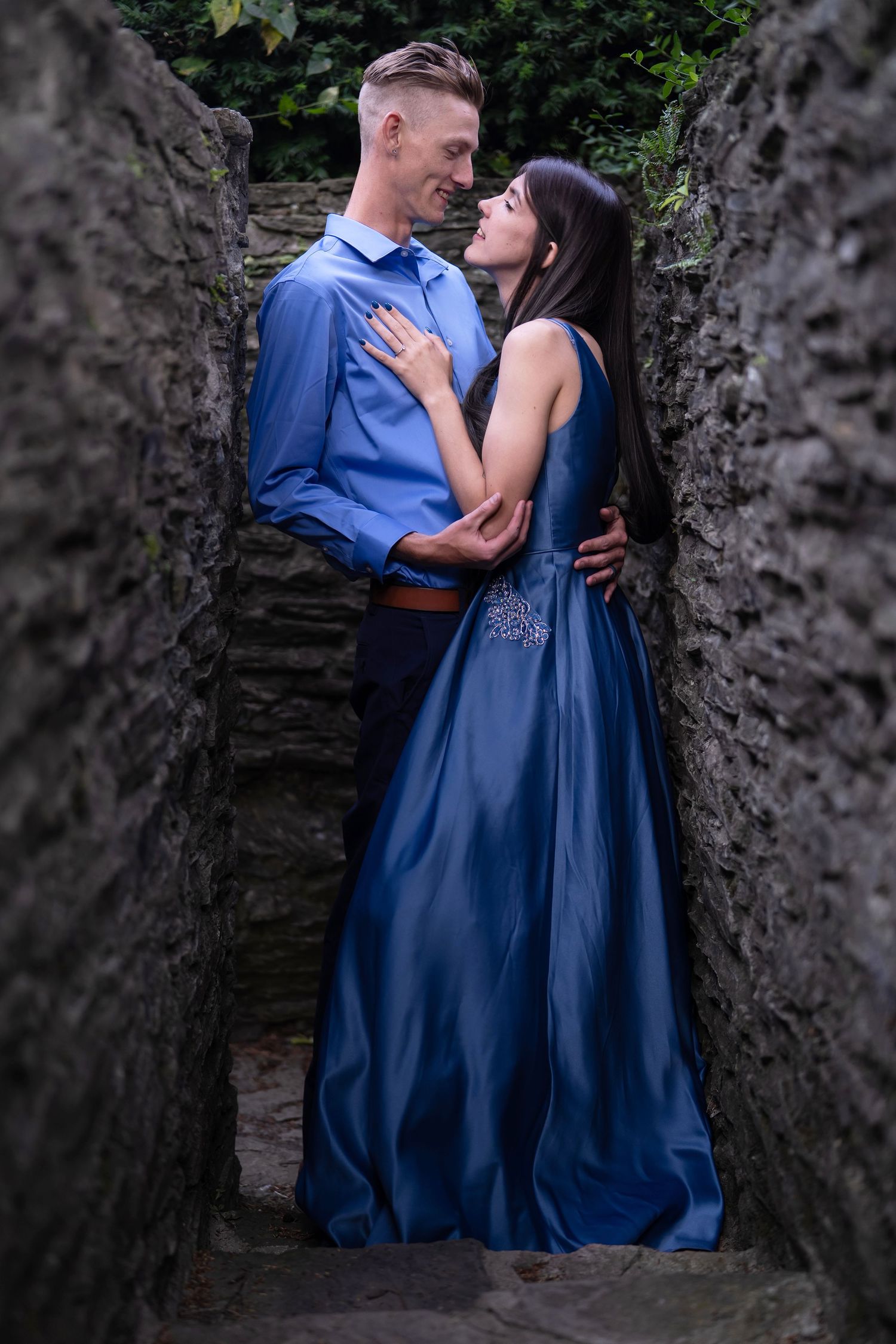 A romantic couple in matching blue formal attire embrace against a stone wall backdrop in an intimate photoshoot.