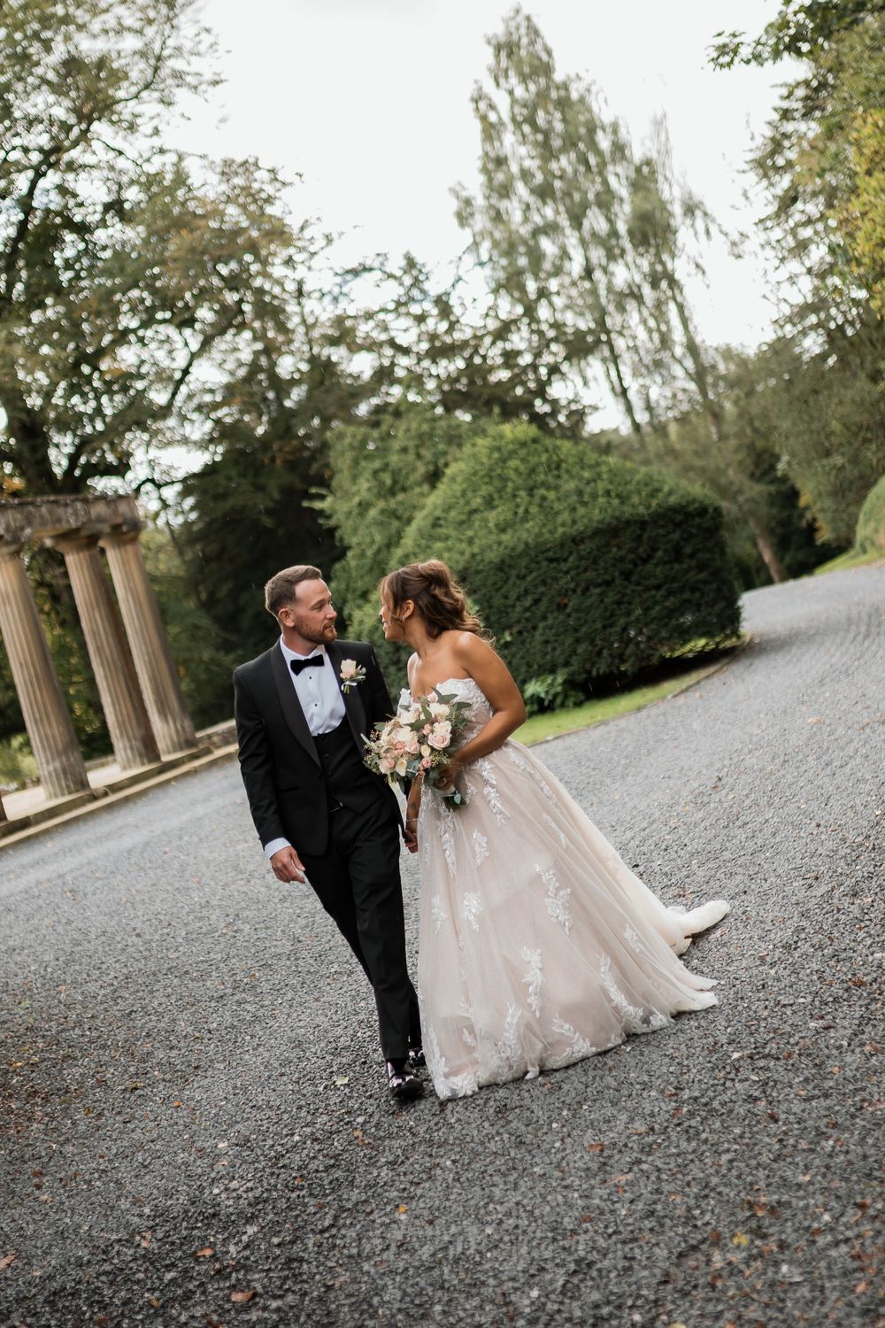 Newlywed couple walking and laughing together on a gravel path lined with trees and columns.