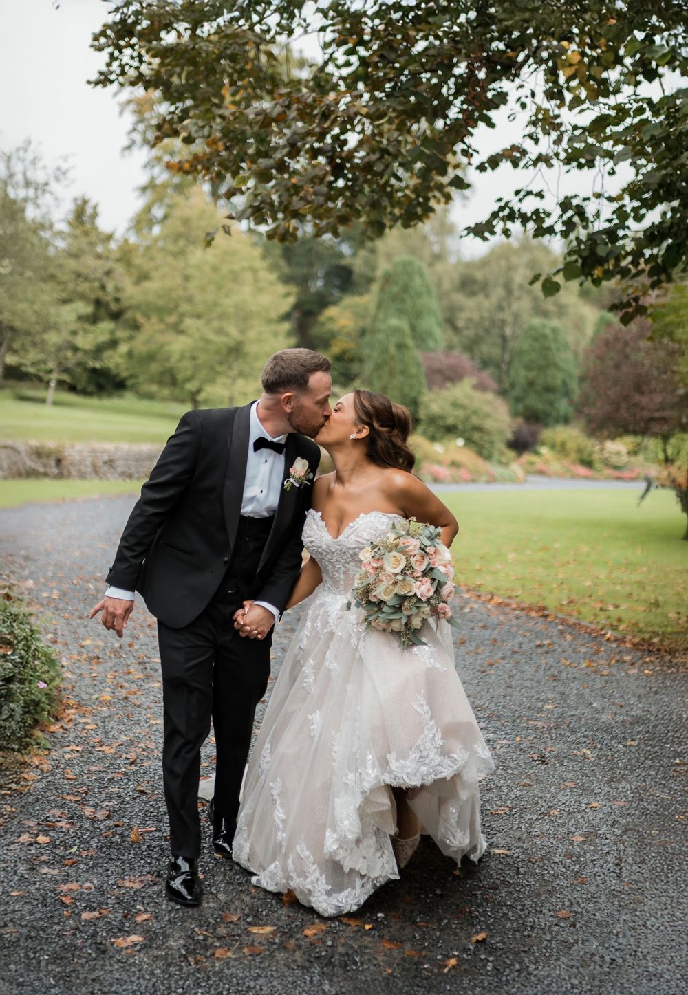Romantic newlywed couple sharing a tender kiss while walking together on a gravel path surrounded by autumn trees.