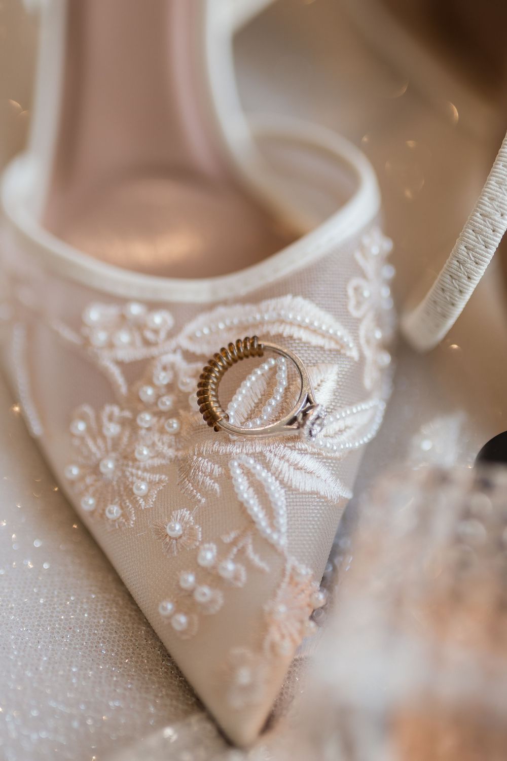 Wedding rings delicately placed on elegant white beaded bridal shoes with intricate embroidery details.