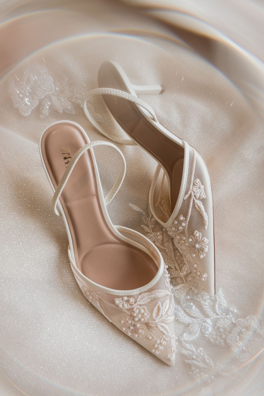 Elegant white lace bridal shoes with delicate floral embroidery displayed on silky fabric background.
