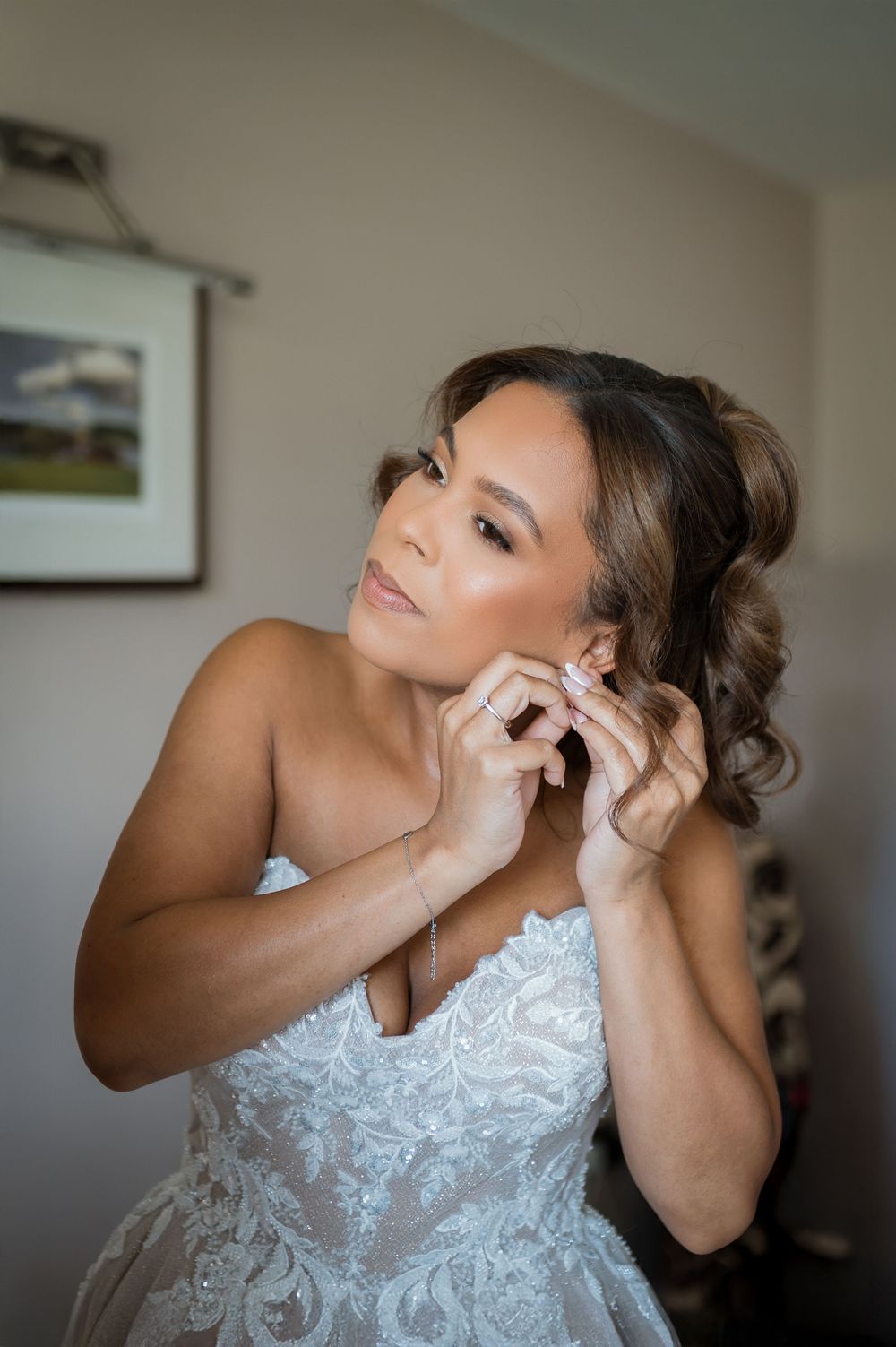 A bride puts on her earrings while getting ready in a delicate white lace wedding dress.