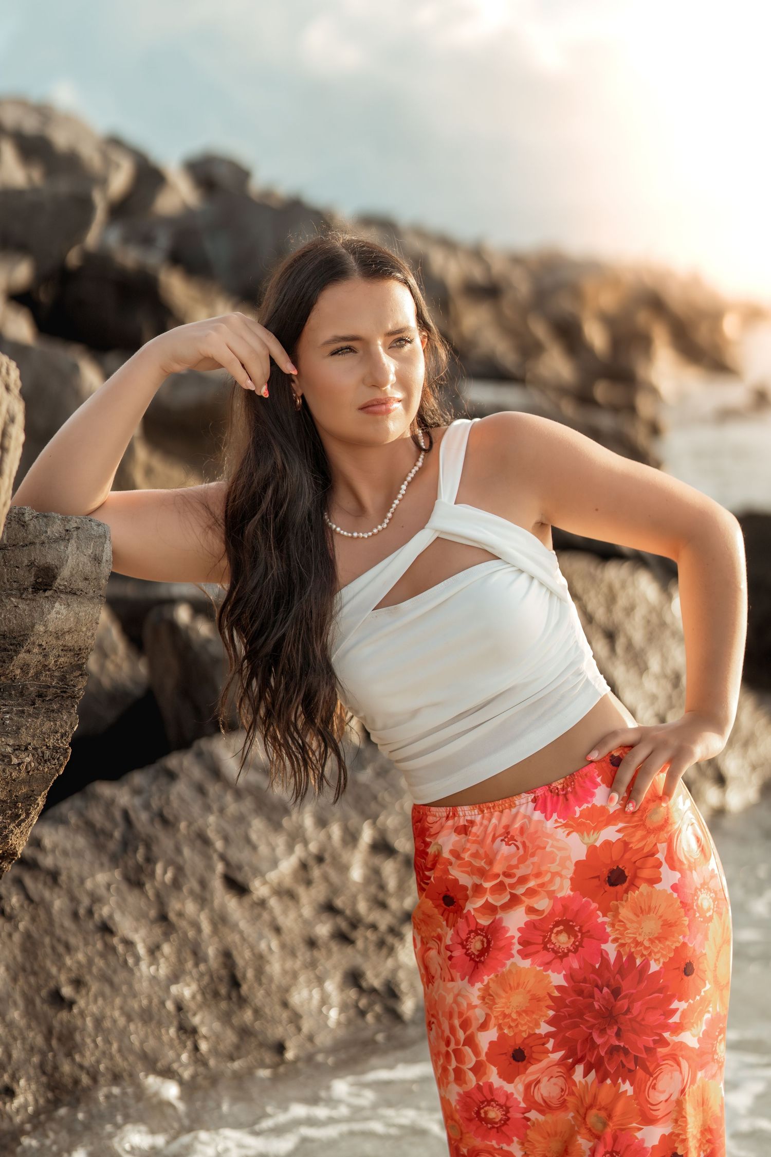 Model in white top and floral skirt poses gracefully against rocky coastal backdrop at sunset.