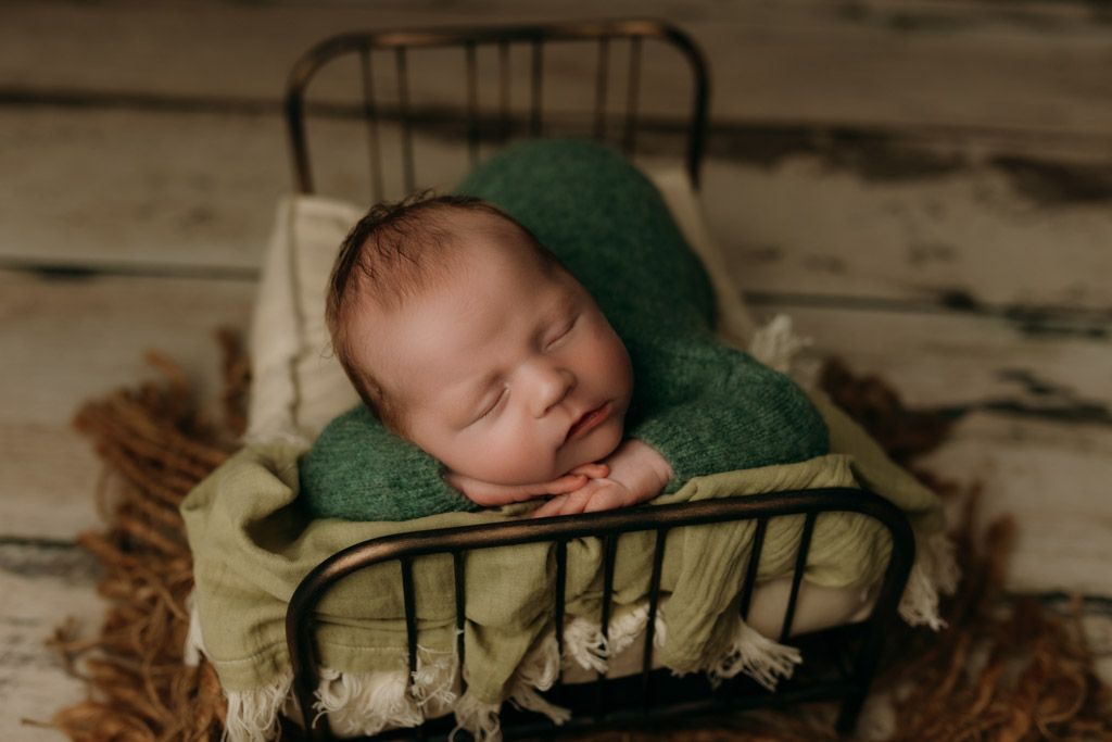 Sleeping baby wearing green bonnet rests peacefully on vintage metal bed with the custom newborn photography setup at studio in victoria bc