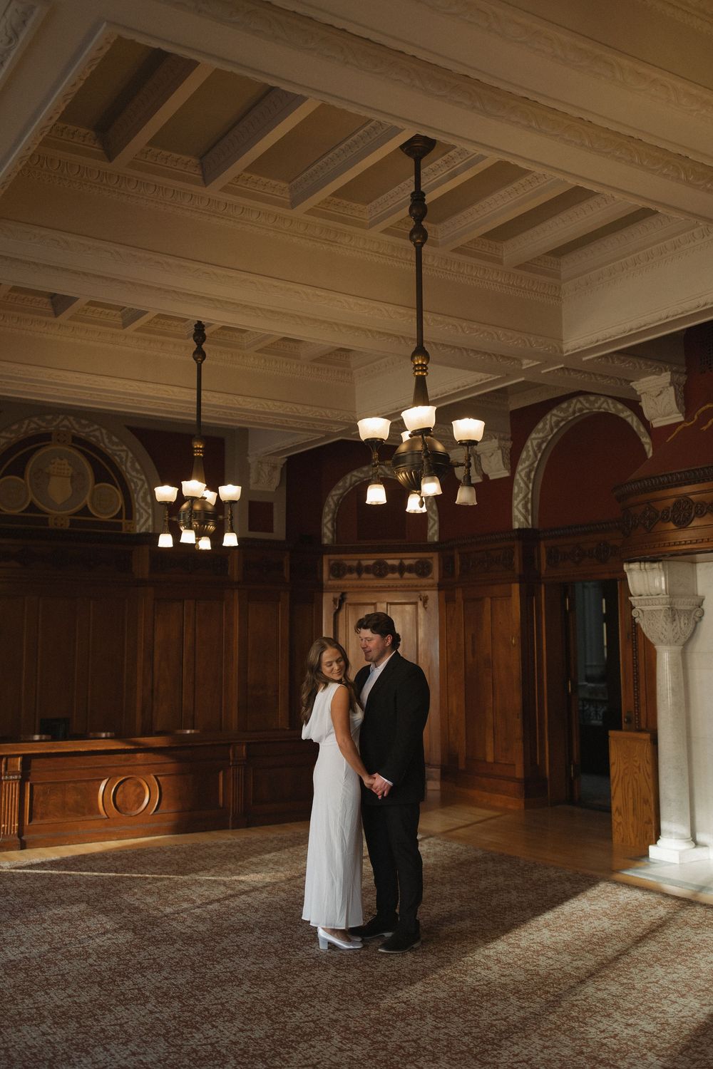 A couple in formal wedding attire stands in an elegant wood-paneled room with ornate chandeliers and columns.