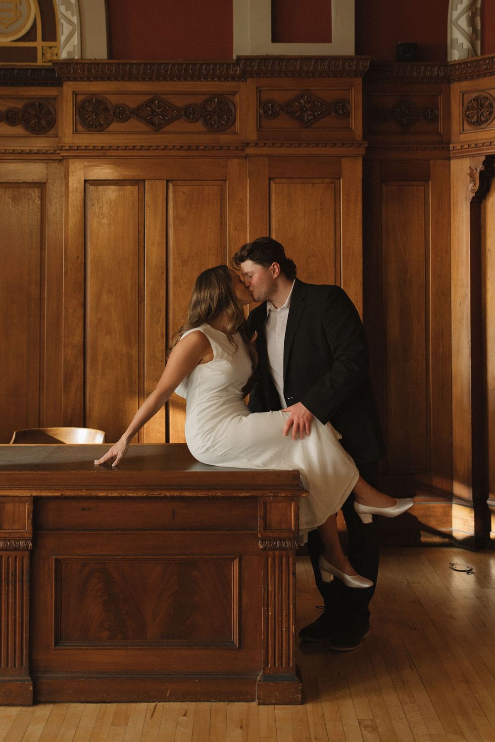 Elegant couple poses on vintage wooden benches in historic interior setting.