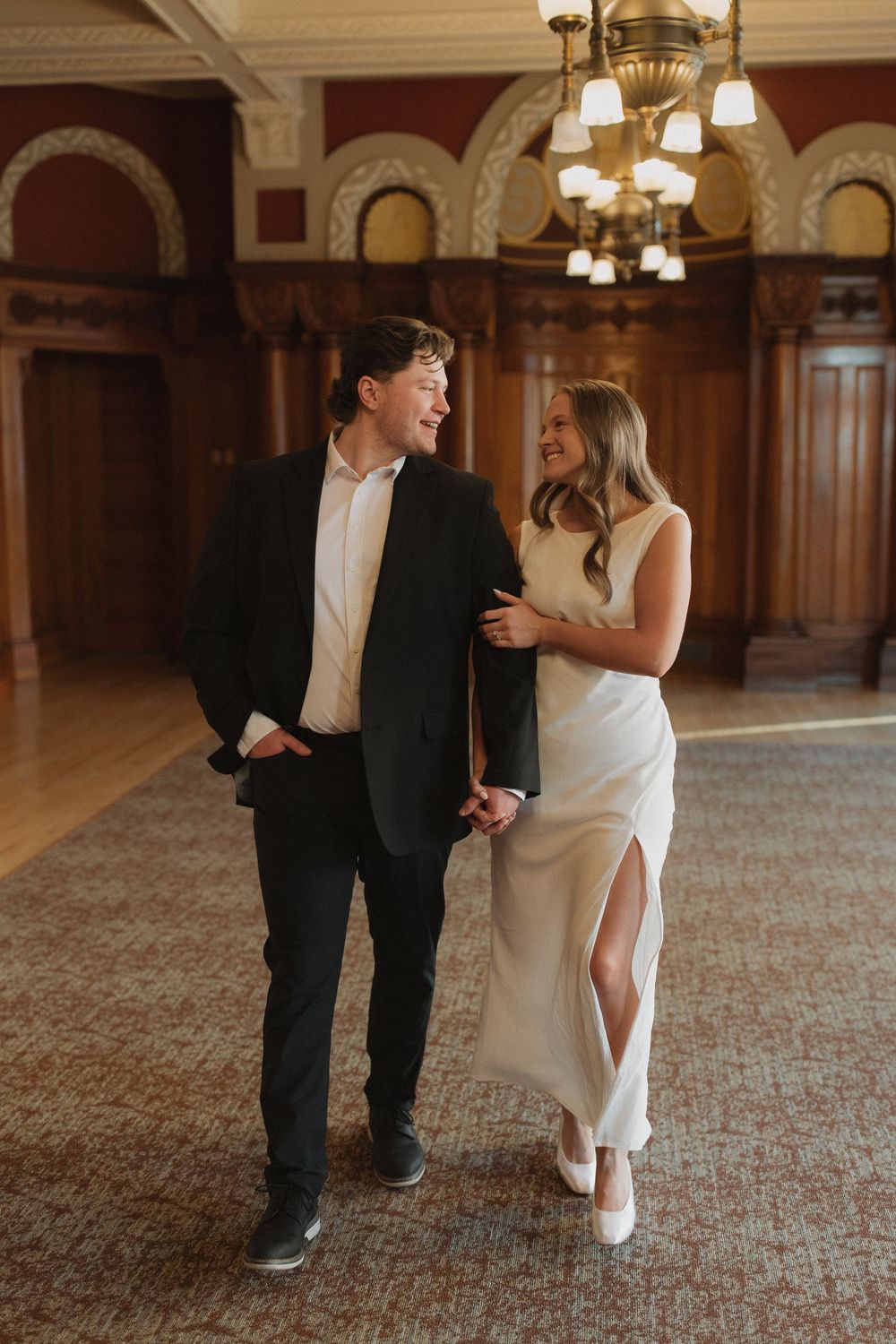 A couple shares a tender moment in an elegant ballroom with chandelier lighting and ornate decor.