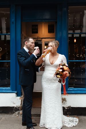 A couple in wedding attire shares a sweet moment while eating sandwiches outside a building with blue trim.