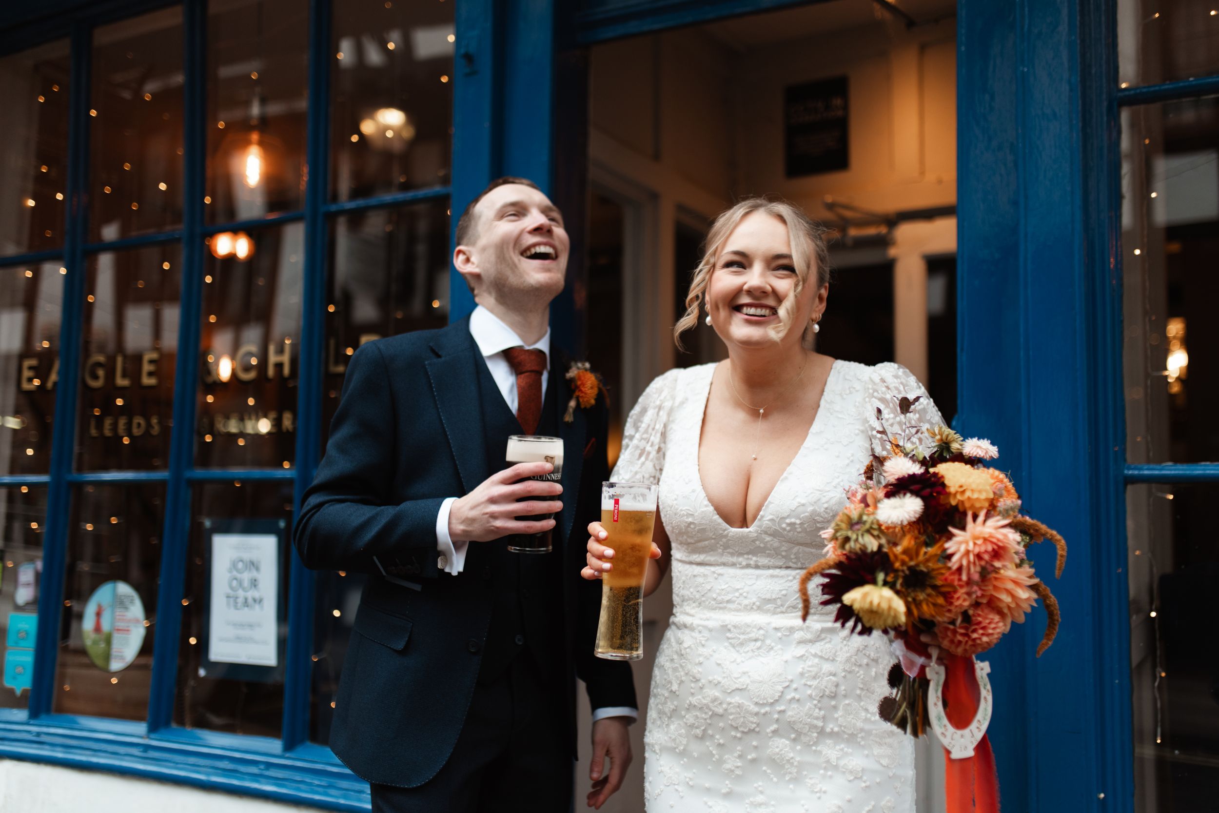 A couple in wedding attire laughs together while holding drinks and a fall-colored bouquet outside a blue storefront.