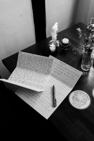 Handwritten letters and notes arranged on a dark table with a candle casting soft light in black and white.