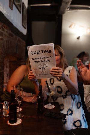 Wedding guests sit at tables with drinks while someone holds a quiz time card during a reception celebration.