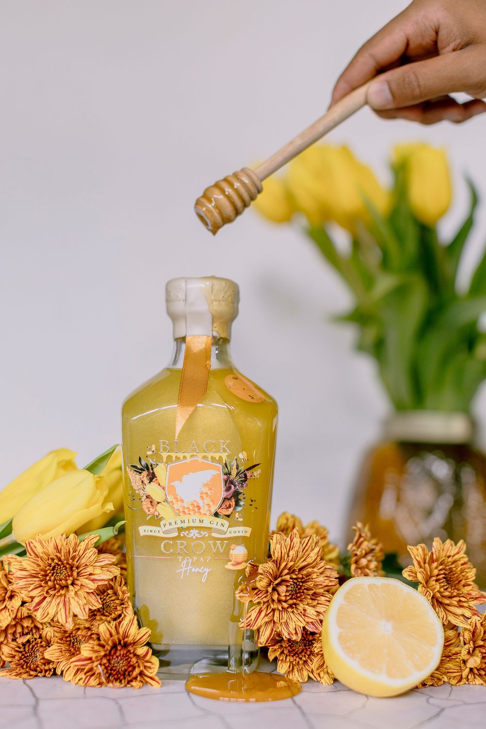 Hand drizzling honey over Black Crow gin bottle with yellow tulips and chrysanthemums on marble.