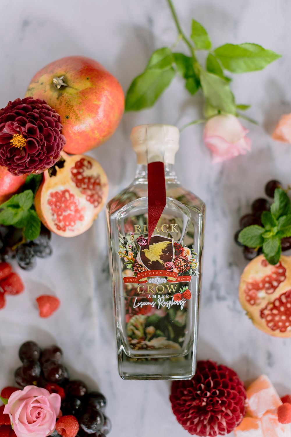 Black Crow Luscious Raspberry gin bottle surrounded by pomegranates, berries, roses, and dahlias.