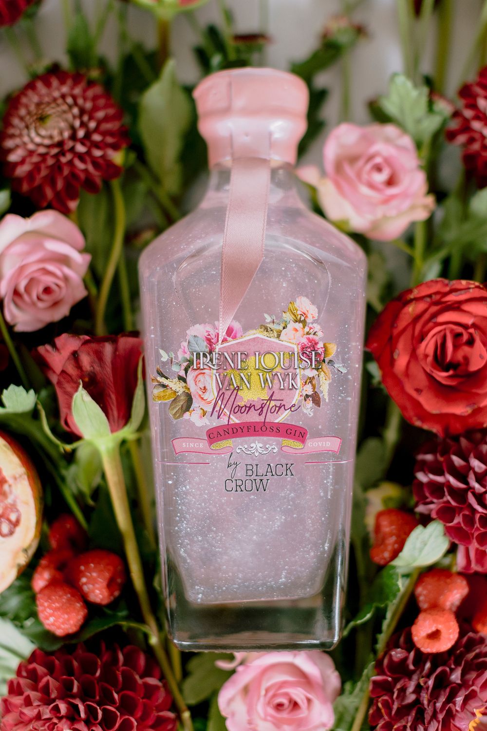 Black Crow Moonstone glitter gin bottle surrounded by pink roses, red dahlias, and raspberries.