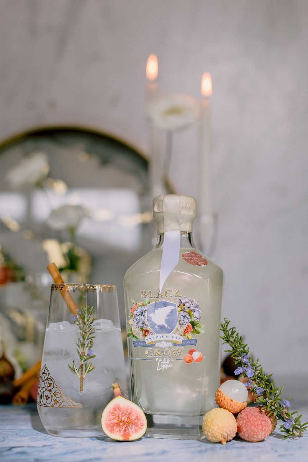 Black Crow Light gin bottle with rosemary cocktail, figs, lychees, and lit candles on marble surface.
