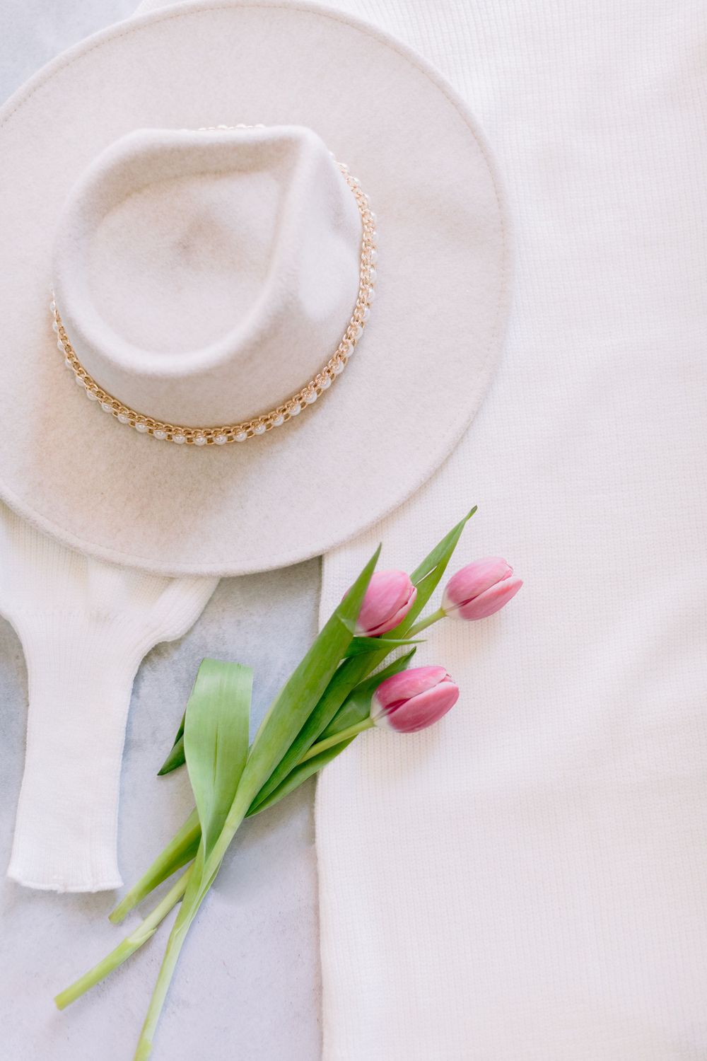White wide-brim felt hat with pearl trim beside pink tulips on white linen fabric, elegant spring flat lay.