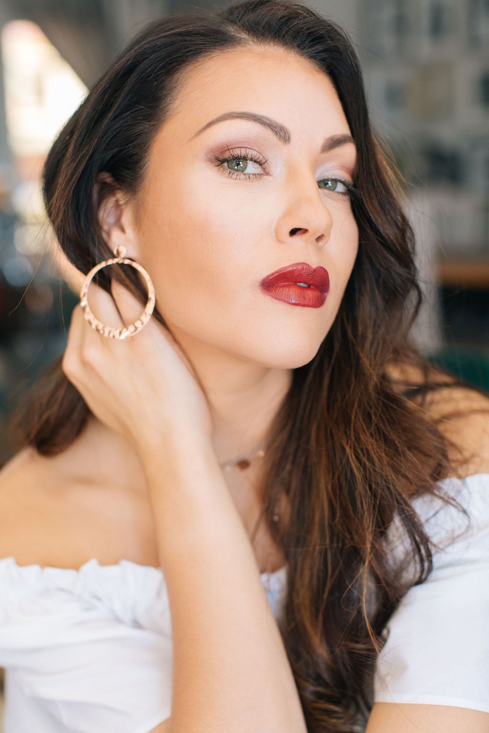 Brunette woman with red lips and gold hoop earrings posing in white top.