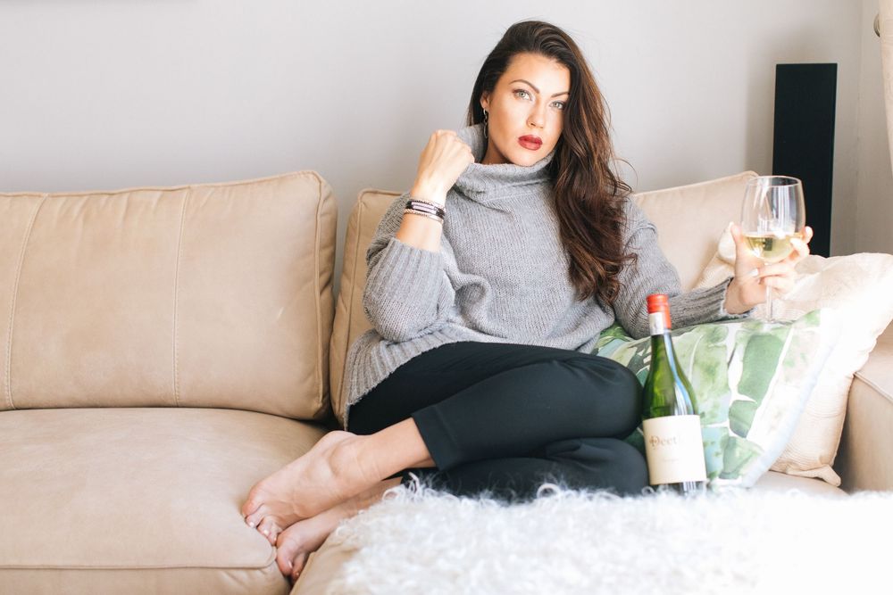 Brunette woman in grey sweater sitting on sofa with white wine bottle.