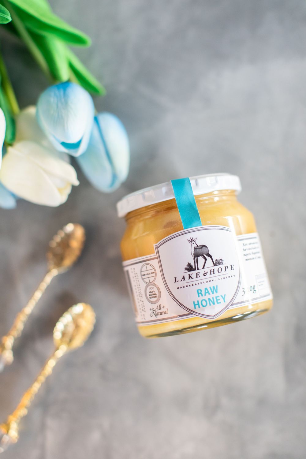 Lake & Hope raw honey jar with golden spoons and blue tulips on grey surface.