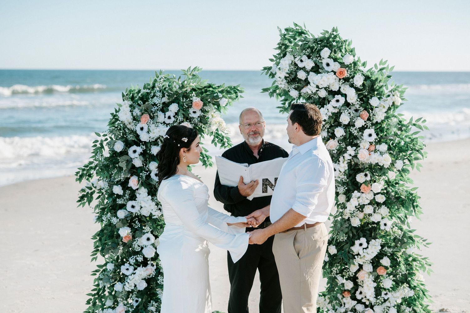 A romantic beach wedding ceremony takes place under a lush green floral arch with ocean waves in the background.