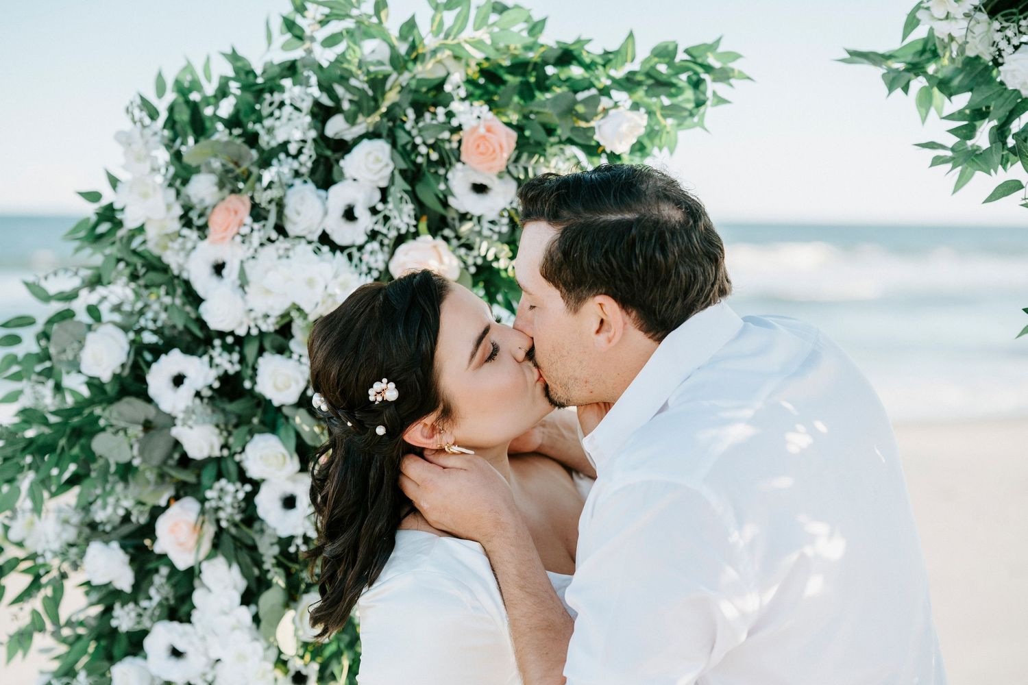 Romantic couple shares a tender kiss under a lush floral arch decorated with white flowers and greenery at a beach wedding.